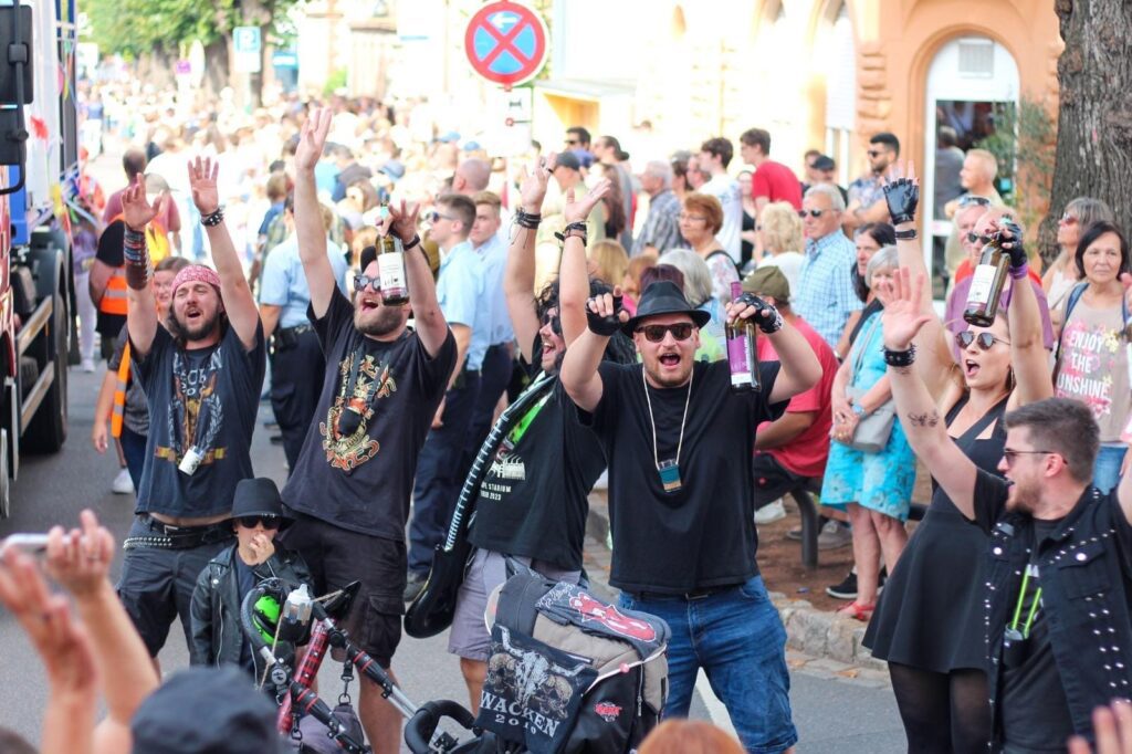 Eine Gruppe schwarz gekleideter Menschen, einige mit Getränken in der Hand, jubeln und heben die Arme während einer lebhaften Partnerstadt-Straßenparade, während im Hintergrund eine große Menschenmenge zusieht.
