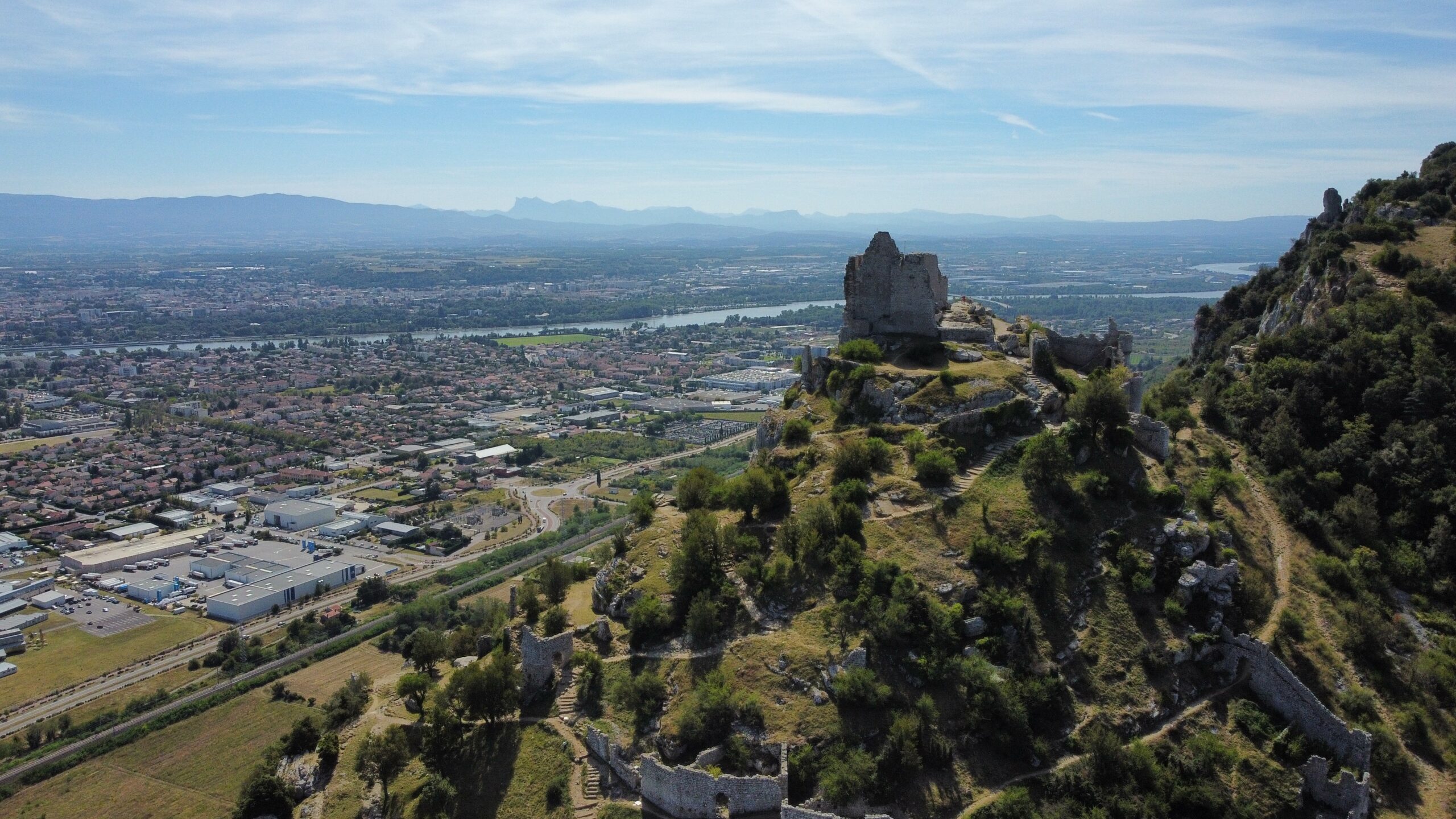Luftaufnahme einer alten Ruine auf einem Hügel mit Blick auf eine ausgedehnte Stadt in Europa, mit einem Fluss, grünen Feldern und fernen Bergen unter einem klaren blauen Himmel.
