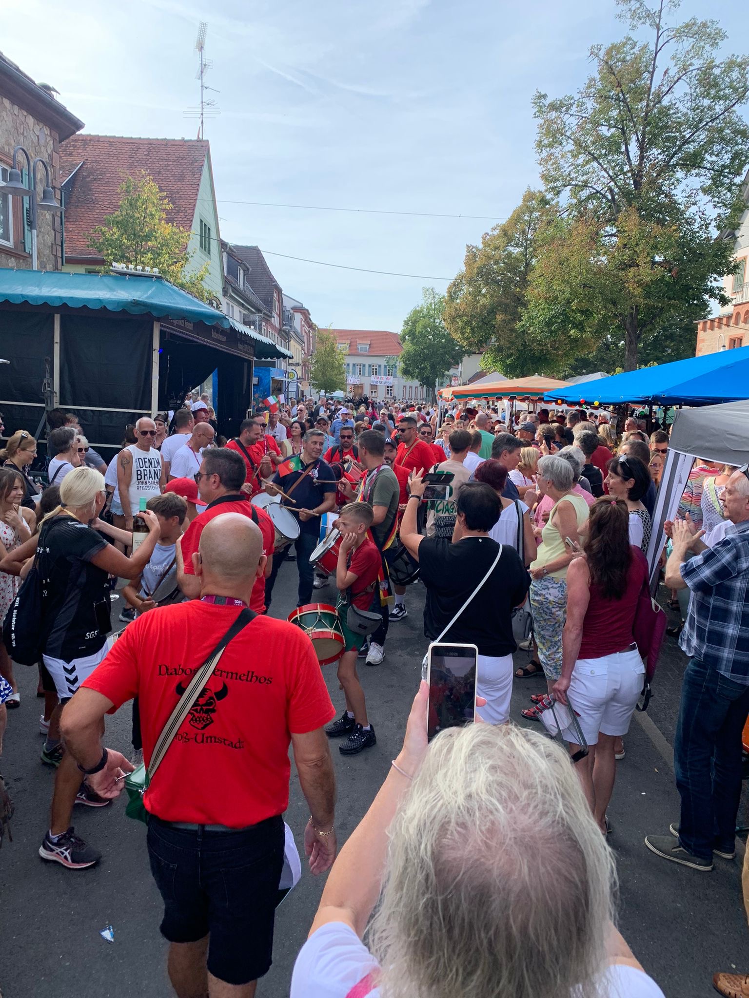 Ein lebhaftes Straßenfest unter freiem Himmel, inspiriert von Europa, mit einer großen Menschenmenge - viele in roten Hemden -, die Musikinstrumente spielt, umgeben von Marktständen, Bäumen und alten Gebäuden unter einem klaren Himmel, um Portugal als Partnerstadt zu feiern.
