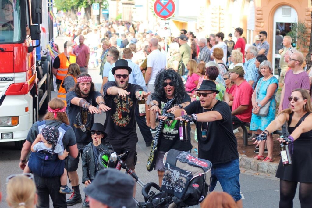 Eine Gruppe in Kostümen, einige mit Perücken und Hüten, steht auf der Straße und imitiert Zombies - vielleicht, um eine Verschwisterung zu feiern. Eine Menschenmenge schaut vom Bürgersteig aus zu, und links ist ein Lastwagen zu sehen.