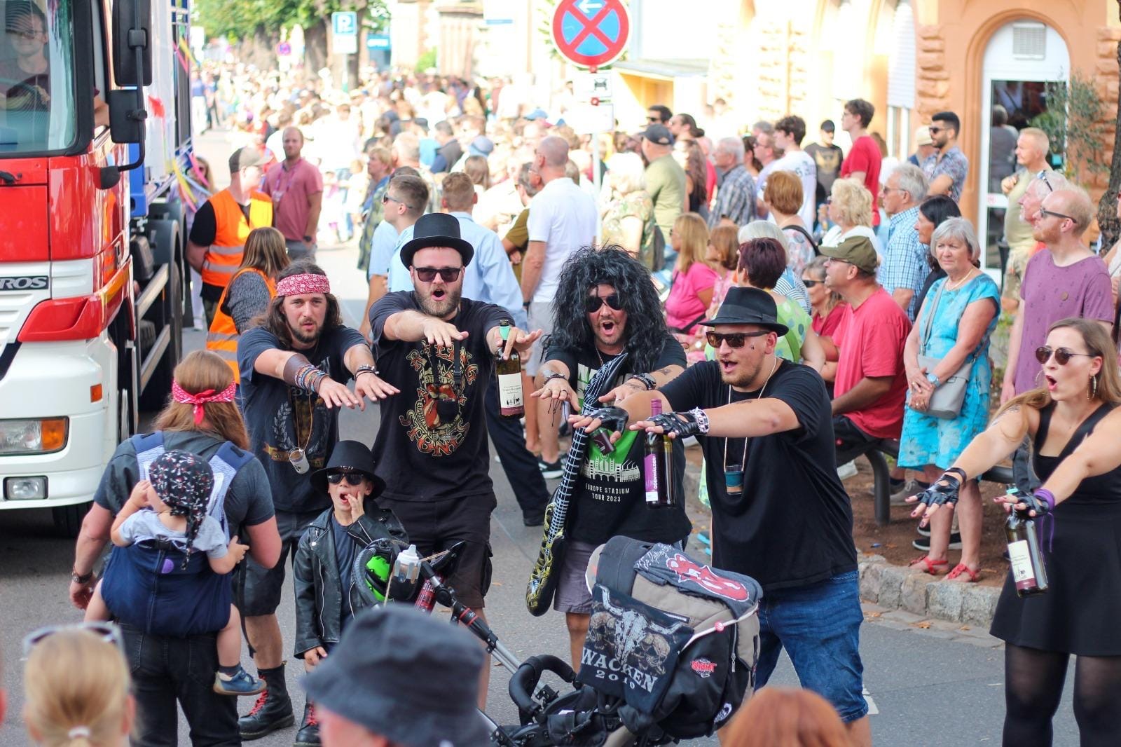 Eine Gruppe in Kostümen, einige mit Perücken und Hüten, steht auf der Straße und imitiert Zombies - vielleicht, um eine Verschwisterung zu feiern. Eine Menschenmenge schaut vom Bürgersteig aus zu, und links ist ein Lastwagen zu sehen.