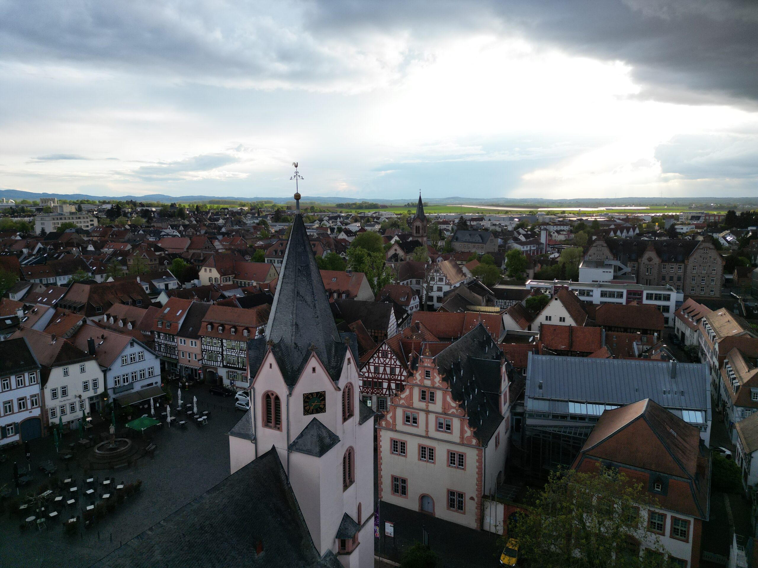 Luftaufnahme von Groß-Umstadt, einer europäischen Stadt mit roten Dächern, einer zentralen Kirche mit Uhrenturm und einem bewölkten Himmel, durch den in der Ferne die Sonne bricht.