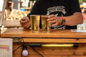 Eine Person stellt zwei Gläser Bier auf einem hölzernen Tresen bei einer São-João-Veranstaltung im Freien ab. Sie trägt ein dunkles T-Shirt und eine Uhr, steht neben einer Speisekarte und einer Glühbirne und fängt die festliche Stimmung des Johannisfestes 2025 in Groß-Umstadt ein.