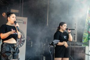 Zwei schwarz gekleidete Frauen treten auf einer Freilichtbühne beim Johannisfest 2025 in Groß-Umstadt auf und singen mit Mikrofonen inmitten von Bühnenausstattung und aufgewirbeltem Rauch. Eine der beiden Frauen gestikuliert ausdrucksstark, während im Hintergrund ein Transparent zu sehen ist.