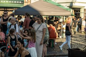 Eine geschäftige Marktszene im Freien mit vielen Menschen, die während des Johannisfestes 2025 in Groß-Umstadt zusammenkommen. Im Vordergrund spielen Kinder miteinander, während sich Erwachsene unter Zelten versammeln, während die Sonne starke Schatten auf den kopfsteingepflasterten Boden wirft.