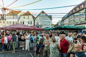Eine große Menschenmenge versammelt sich beim Johannisfest 2025 auf dem Stadtplatz von Groß-Umstadt, umgeben von Fachwerkhäusern, Sonnenschirmen und bunten Luftschlangen. Menschen aller Altersgruppen unterhalten sich und genießen die lebhafte Festatmosphäre.
