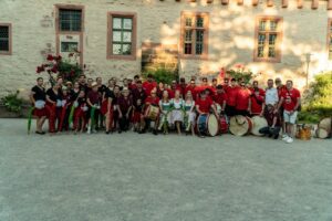 Eine große Gruppe von Menschen, einige mit Trommeln in der Hand und andere in farbenfrohen Kostümen, posieren während des Johannisfestes 2025 in Groß-Umstadt gemeinsam im Freien vor einem Steingebäude mit Fenstern und Blumensträuchern.