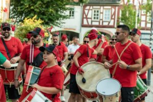 Eine Gruppe von Menschen mit roten Hemden und Hüten trommelt tagsüber auf dem Johannisfest 2025 in Groß-Umstadt im Freien. Bäume und Fachwerkhäuser bilden eine festliche Kulisse, die an die Feste von São João erinnert.