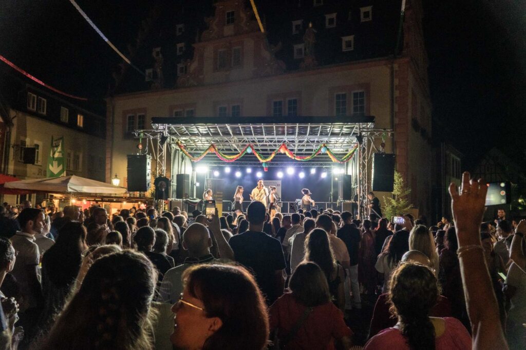 Eine große Menschenmenge verfolgt den Auftritt einer Live-Band auf einer nächtlichen Freilichtbühne mit bunten Lichtern und Dekorationen über der Bühne. Die Veranstaltung findet während des Johannisfestes 2025 in Groß-Umstadt vor einem historischen Gebäude statt.