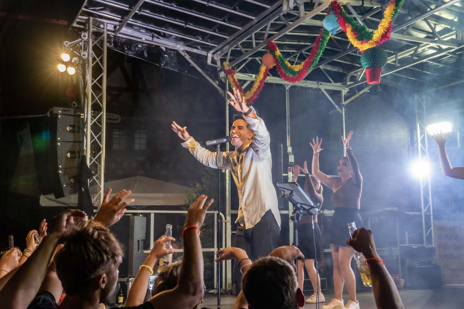 Ein Sänger im weißen Hemd singt energisch auf einer nächtlichen Freilichtbühne während des Johannisfestes 2025, begleitet von Backgroundtänzern und bunten Dekorationen. Das Publikum jubelt mit erhobenen Händen und Flaschen, Lichter erhellen die lebhafte Szene.