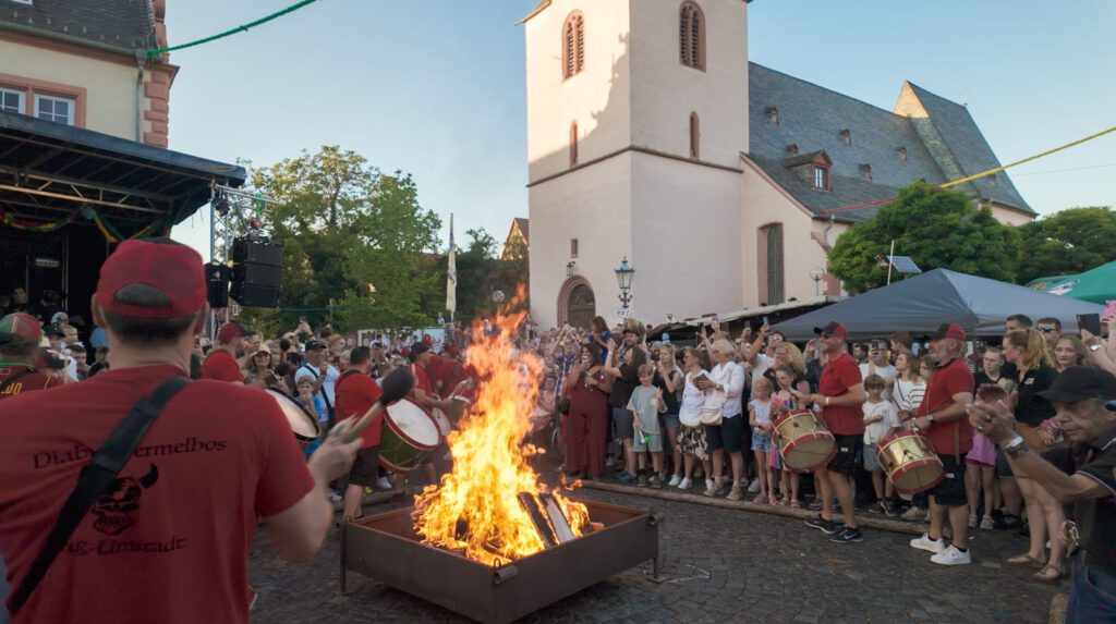 Eine lebhafte Menschenmenge versammelt sich auf dem Marktplatz von Groß-Umstadt in der Nähe der Kirche und beobachtet die Darsteller in roten Hemden, die während des Johannisfestes 2025 um ein großes offenes Feuer herum trommeln.