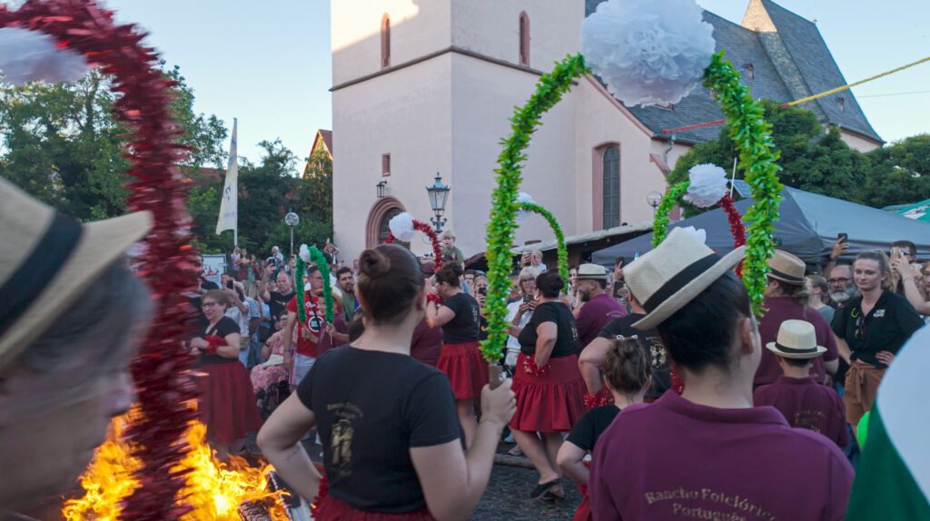 Eine Gruppe von Menschen in traditioneller Kleidung tanzt auf dem Johannisfest 2025 in Groß-Umstadt und hält grüne Bögen mit weißen Bommeln in der Nähe einer Kirche. Im Vordergrund brennt ein kleines Feuer, während die Menge dieses festliche São João-Fest genießt.