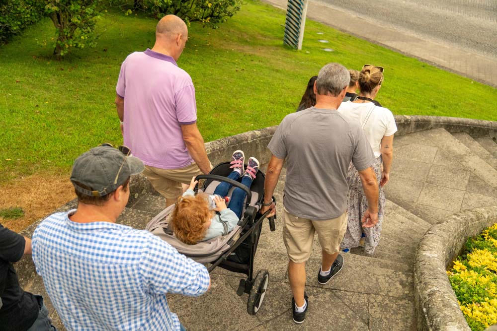 Eine Gruppe Erwachsener geht mit einem Kinderwagen, in dem ein kleines Kind liegt, eine geschwungene Steintreppe hinunter. Die grüne, gut gepflegte Fläche lässt auf einen Park oder Garten schließen - vielleicht während des Festes São Bento in Santo Tirso, 2025.