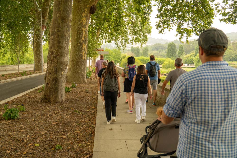 Eine Gruppe von Menschen, darunter Erwachsene und Kinder, geht gemeinsam einen von Bäumen gesäumten Weg neben einem Park in Santo Tirso entlang. Eine Person schiebt einen Kinderwagen, während sie das üppige Grün genießen und den Geist des Festes São Bento 2025 zelebrieren.