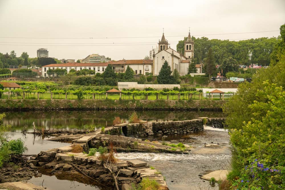 Ein Fluss mit einem Steinwehr fließt durch die grüne Landschaft von Santo Tirso. Im Hintergrund, in der Nähe der Festung São Bento 2025, steht eine historische weiße Kirche mit zwei Glockentürmen inmitten von Bäumen und einem bewölkten Himmel.