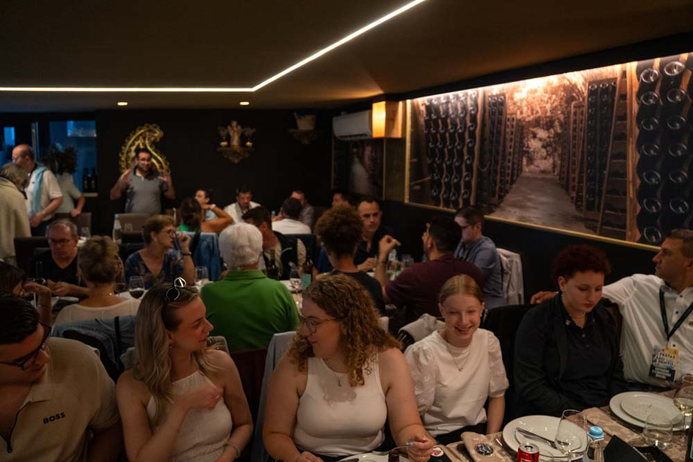 Eine große Delegation sitzt an einem langen Esstisch in einem schwach beleuchteten Restaurant in Santo Tirso, unterhält sich und genießt eine Mahlzeit. An der Wand stehen Weinflaschen, und die Atmosphäre wirkt lebendig und gesellig und erinnert an das Fest São Bento 2025.