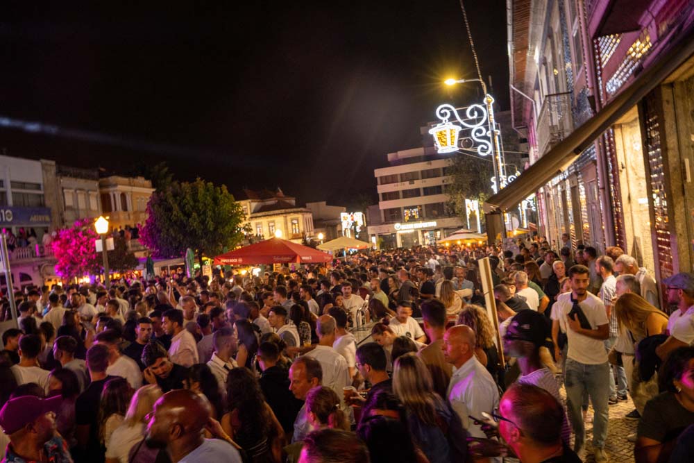 Eine große Menschenmenge versammelt sich nachts im Freien zum lebhaften Fest São Bento 2025 in Santo Tirso, umgeben von Leuchtreklamen, Essensständen und dekorativen Lichtern. Die Atmosphäre ist festlich und energiegeladen, als sich die Delegation den Feierlichkeiten anschließt.