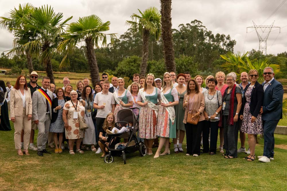 Eine große Gruppe, Teil der São Bento 2025 Delegation aus Santo Tirso, posiert im Freien auf einer Wiese mit Palmen im Hintergrund; mehrere Frauen tragen traditionelle Kleider, und in der vorderen Mitte ist ein Baby in einem Kinderwagen zu sehen.