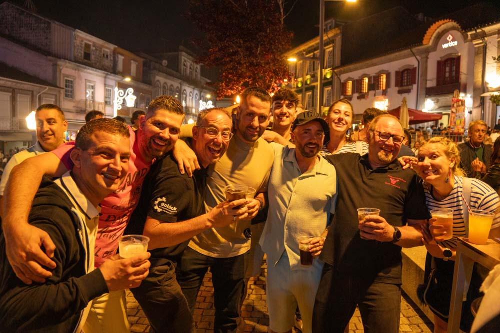 Eine Gruppe lächelnder Menschen mit Getränken in der Hand posiert nachts auf einer belebten, gut beleuchteten Straße in Santo Tirso während des Festes São Bento 2025, mit historischen Gebäuden im Hintergrund. Die Szene vermittelt eine festliche und fröhliche Atmosphäre.