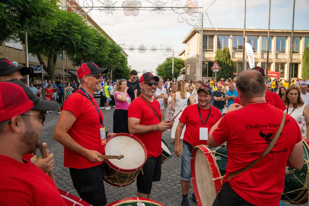 Eine Gruppe lächelnder Musiker in passenden roten Hemden und Hüten trommelt auf dem Fest São Bento 2025 in Santo Tirso, umgeben von einer Menge Schaulustiger und festlicher Dekoration über dem Kopf.