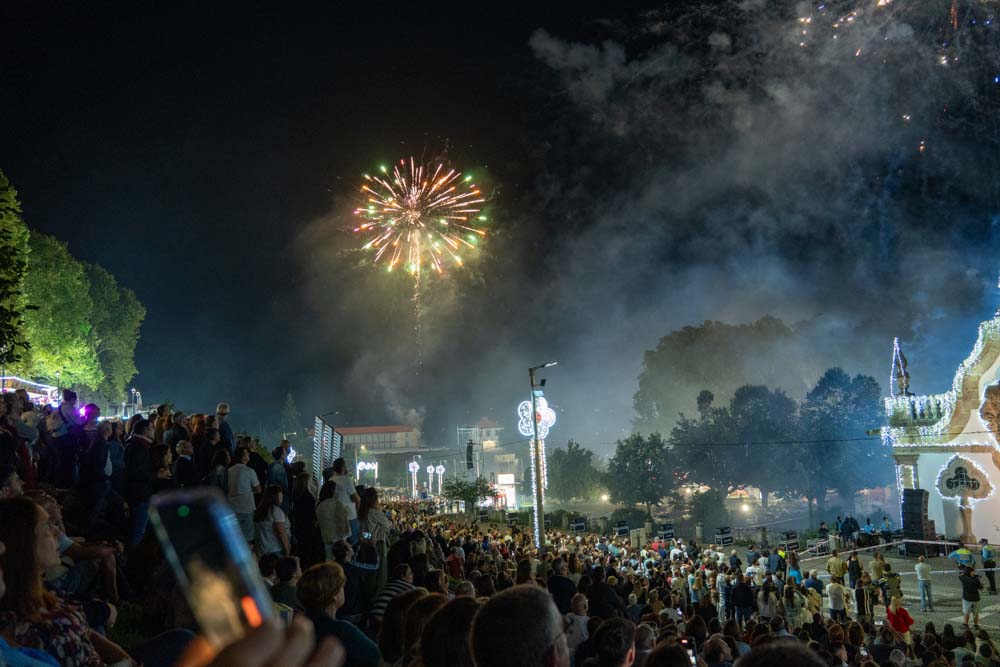 Während des Festes São Bento 2025 in Santo Tirso versammelt sich nachts eine große Menschenmenge im Freien, um das bunte Feuerwerk zu beobachten, das den Himmel erhellt, mit Bäumen und beleuchteten Gebäuden im Hintergrund. Einige Leute machen Fotos mit ihren Handys.