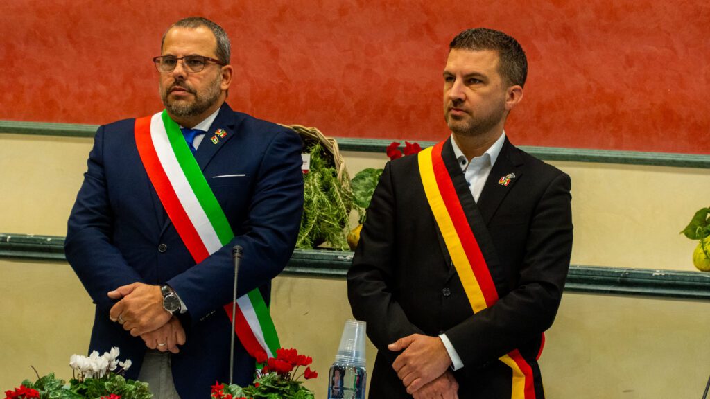 Zwei Männer in formellen Anzügen stehen drinnen, jeder trägt eine Schärpe - eine in den Farben der italienischen Flagge, die für Dicomano und seine Fiera d'Ottobre steht, die andere in Schwarz, Gelb und Rot für ihre Partnerstadt. Auf dem Tisch stehen Topfblumen und Glaswaren.