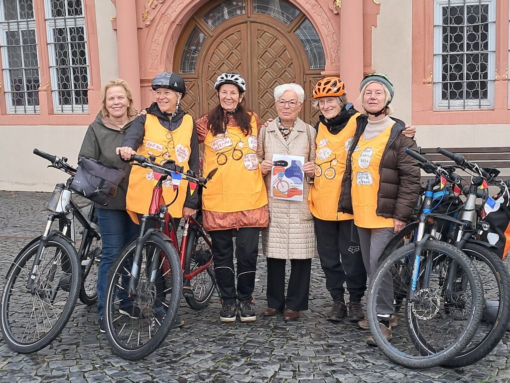 Fünf Frauen mit Helmen und gelben Westen stehen mit ihren Rädern neben einer älteren Frau, die ein Büchlein in der Hand hält, und posieren vor einem großen, verzierten Holztor - bereit für die Tour d'Amitié 2024 mit ihren französischen Freunden.