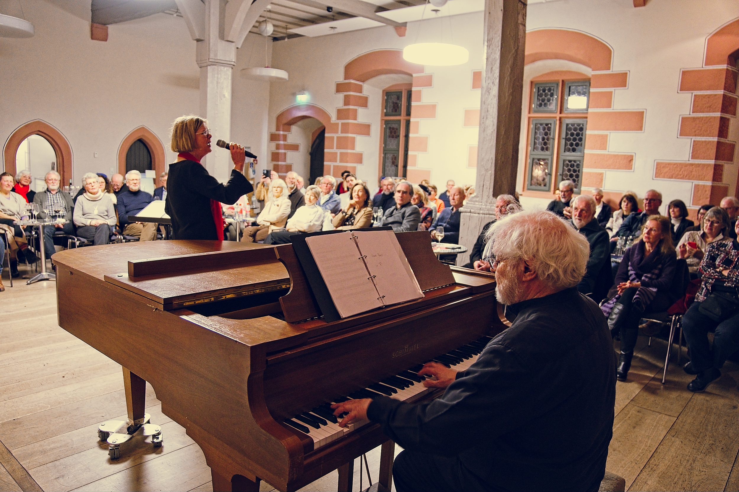 Eine Frau singt ein gefühlvolles Chanson in ein Mikrofon, während ein Mann in einem geräumigen Raum mit gewölbten Fenstern einen Flügel spielt. Das Publikum sitzt aufmerksam da und lässt sich von der warmen, intimen Atmosphäre und der Geschichte der Freundschaft, die sich durch die Musik entfaltet, mitreißen.
