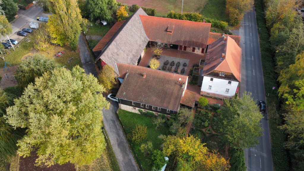 Luftaufnahme eines Gebäudekomplexes mit roten Ziegeldächern, umgeben von Bäumen und Grünflächen, angrenzend an eine Straße mit parkenden Autos. Wege und ein kleiner Innenhof deuten auf die Vorbereitungen für das bevorstehende Internationale Sommerfest hin.