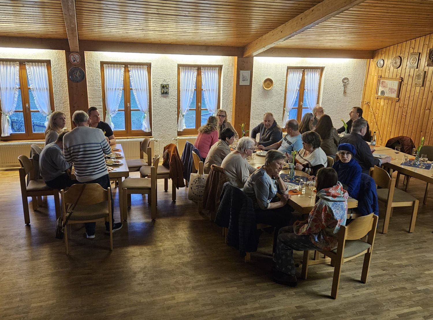 Eine Gruppe von Menschen sitzt um zwei lange Tische in einem gemütlichen Holzzimmer und isst, trinkt und unterhält sich bei einem freundlichen Helferabend. Natürliches Licht fällt durch die mit Vorhängen versehenen Fenster und trägt zu der ungezwungenen und einladenden Atmosphäre bei.